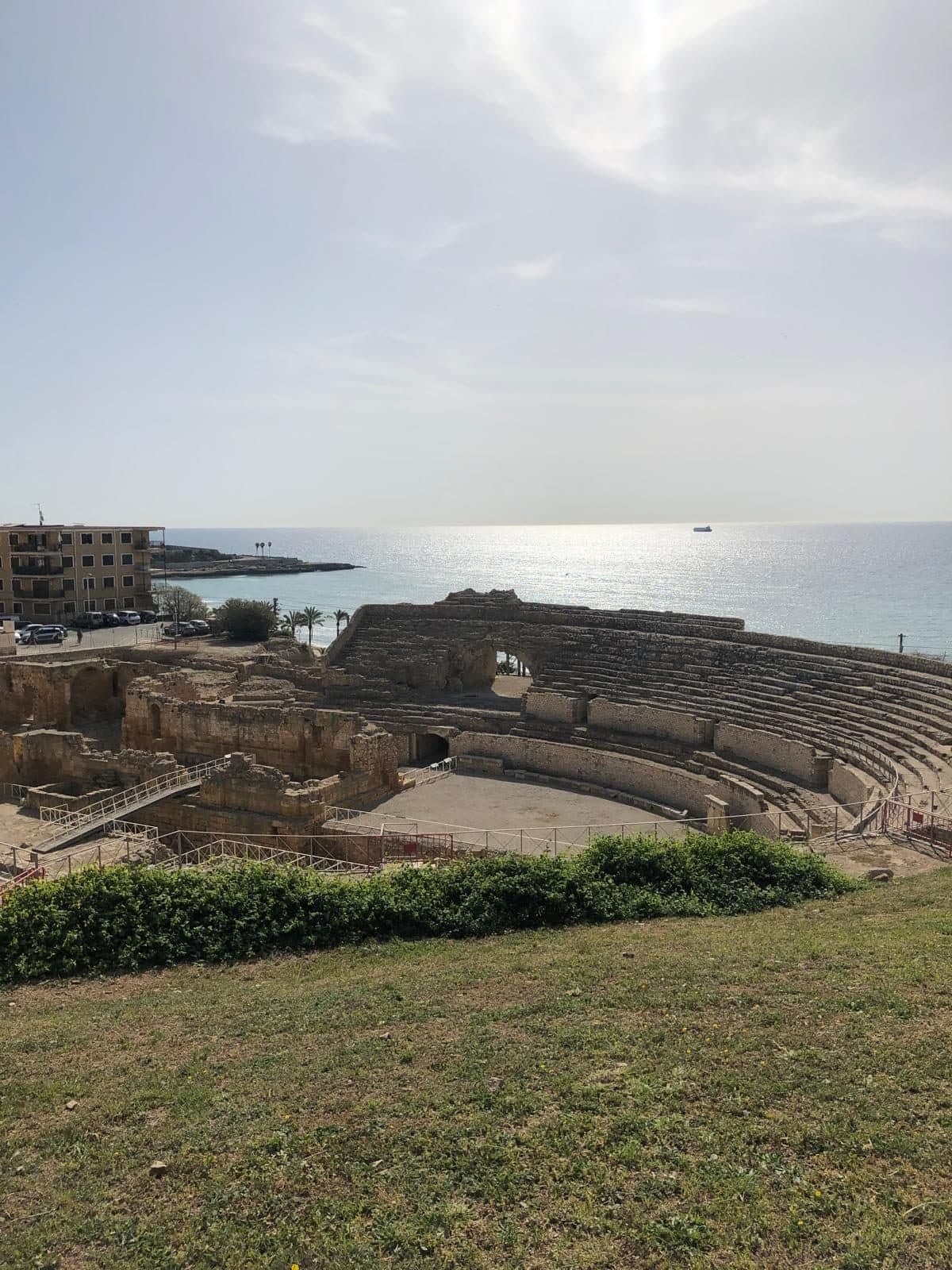 The Roman amphitheatre of Tarraco seen from a grassy bank above, the sea beyond