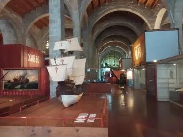 Una petita embarcació blanca exposada davant el mur vermell de la sala